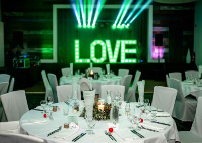 Festsaal mit rund gedeckten Tischen, weißen Stuhlhussen und großem grünen LED-Schriftzug 'LOVE' an der Wand, darüber grüne Lichtstrahlen.