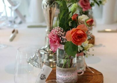 Tischdekoration mit silbernem Kerzenhalter, Glasvase mit orangefarbenen und rosa Blumen auf Holzscheibe, Gläser und weiße Tischdecke.