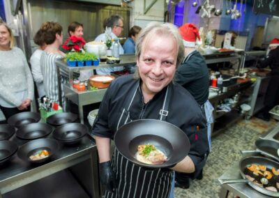 Koch mit grauen Haaren und schwarzer Schürze hält eine schwarze Pfanne mit angerichtetem Gericht in einer geschäftigen Küche mit mehreren schwarzen Pfannen und Personen im Hintergrund.