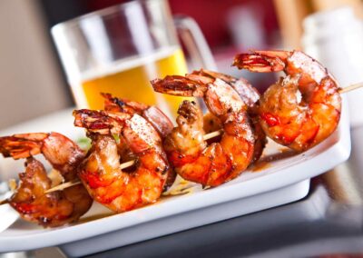 Fünf gegrillte Garnelen auf einem Holzspieß, serviert auf einem weißen Teller, im Hintergrund ein Glas Bier.