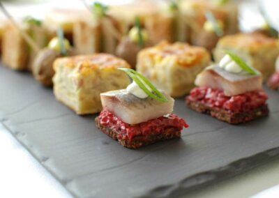 Mehrere kleine Häppchen auf einer schwarzen Steinplatte, darunter ein Stück Fisch auf rotem Belag und ein quadratisches Gebäck mit grüner Garnitur.