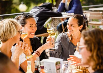Mehrere Personen sitzen an einem Tisch im Freien und stoßen mit Weingläsern an.