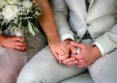 Braut und Bräutigam sitzen nebeneinander, halten sich an den Händen, Braut hält einen Blumenstrauß mit weißen Rosen und Schleierkraut.