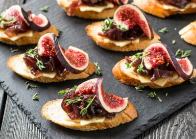 Mehrere geröstete Baguettescheiben mit Käse, Feigenstücken, dunkler Marmelade und Thymianzweigen auf dunklem Untergrund.