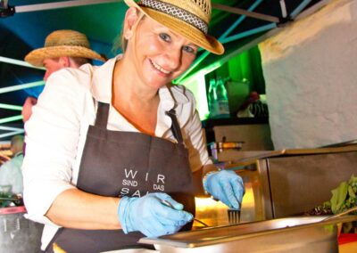 Service-Mitarbeiterin mit Strohhut und schwarzer Schürze bereitet Speisen unter grünem Zelt zu.