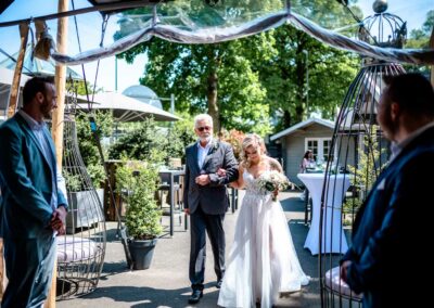 Braut in weißem Kleid mit Blumenstrauß geht Arm in Arm mit Mann im Anzug durch einen mit Pflanzen dekorierten Außenbereich, zwei Männer in Anzügen stehen links und rechts.