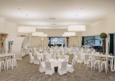 Großer Festsaal mit mehreren runden Tischen, weiß gedeckt und mit weißen Stuhlhussen, helle Lampen an der Decke, Holzboden und großen Fenstern.