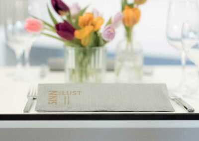 Tisch im Festsaal mit grauem Menü, Besteck und Gläsern, dahinter Vasen mit bunten Tulpen in Gelb, Lila und Rosa.