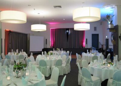 Festsaal mit runden, weiß eingedeckten Tischen und Stühlen, großer Bühne mit Vorhang und rosa Beleuchtung, hängende Lampen.