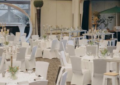 Festsaal mit mehreren runden Tischen, weiß bezogenen Stühlen, großen weißen Deckenlampen und Holzboden.