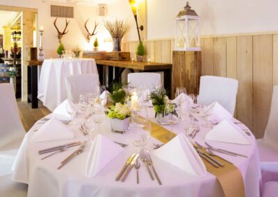 Rund gedeckter Tisch mit weißer Tischdecke, gefalteten Servietten, Besteck, Gläsern und grünen Pflanzen in einem hellen Raum mit Holzwandverkleidung und Hirschgeweih an der Wand.