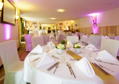 Festlich gedeckte runde Tische mit weißen Hussen, gefalteten Servietten und Gläsern in einem hellen Landhausstubenraum mit Hirschgeweihen an den Wänden.