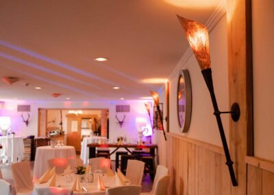 Landhausstube mit gedeckten Tischen, weißen Stühlen, Wandlampen mit warmem Licht und Holzwandverkleidung.