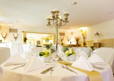 Rund gedeckter Tisch mit weißen Stoffservietten, Gläsern, Besteck und Blumenarrangements in einer hellen Landhausstube mit Hirschgeweihen an den Wänden.