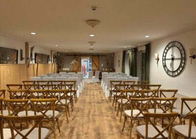 Landhausstube mit Holzboden, mehreren Reihen Holzbänke mit weißen Sitzpolstern, großer runder Wanduhr und Fenster mit grünen Vorhängen.