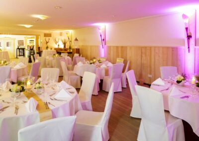 Festlich gedeckte runde Tische mit weißen Hussen, Kerzen und Blumenarrangements in einem hellen Raum mit Holzvertäfelung und Hirschgeweihen an der Wand.