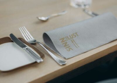 Tisch mit grauer Stoffserviette mit goldener Aufschrift, Besteck und leerem Wasserglas auf hellem Holz.
