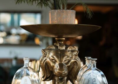 Goldfarbener Löwenkopf als Dekoration auf einem Sockel, darüber eine Pflanze in einem geriffelten Glasgefäß, flankiert von zwei klaren Flaschen mit weißen Etiketten.