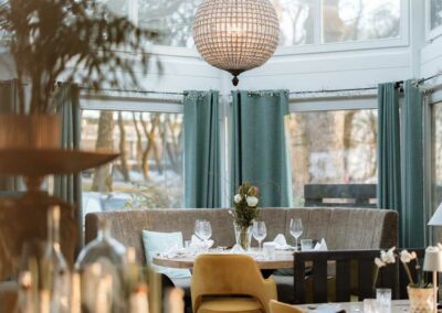 Essbereich mit runden Tischen, gelben Stühlen, grauer Sitzbank, großen Fenstern und einer runden, gläsernen Deckenleuchte.