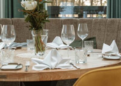 Runder Holztisch mit weiß gefalteten Servietten, mehreren Gläsern und einer Vase mit Blumen, im Hintergrund Fenster mit Blick auf Bäume.