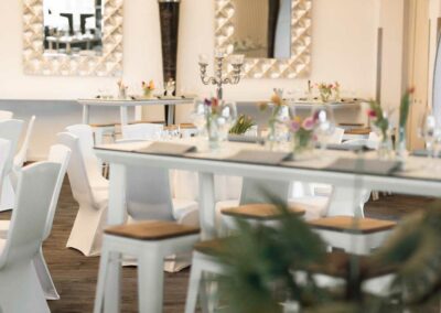 Moderner Gastraum mit hohen weißen Tischen und Hockern, zwei rechteckigen Spiegeln mit geometrischem Rahmen und großen runden Deckenlampen.