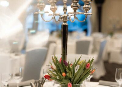 Silberner Kerzenhalter mit fünf Armen auf einem Tisch, davor Glasvase mit roten und orangefarbenen Tulpen, gedeckter Tisch im Hintergrund.