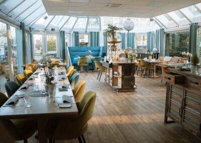 Helles Restaurant mit Holzboden, mehreren Tischen, grünen und gelben Stühlen, blauer Sitzbank und großen Fenstern mit blauen Vorhängen.