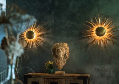 Zwei runde Wandlampen mit flammenartigem Lichtmuster und eine Löwenkopf-Skulptur auf einem Holztisch vor dunkler Wand.