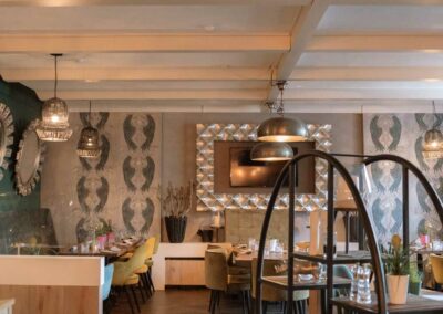 Innenraum eines Restaurants mit Holzboden, mehreren Tischen und Stühlen, dekorativen Lampen und Spiegeln an den Wänden.