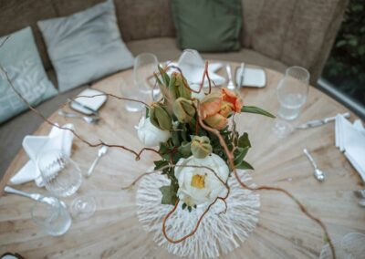 Runder Holztisch mit Blumenstrauß aus weißen und orangefarbenen Blüten, umgeben von Gläsern, Besteck und Servietten.