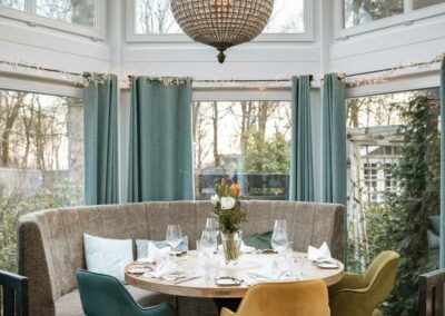 Runder Esstisch mit weißem Marmor und gedecktem Geschirr, umgeben von gepolsterten Stühlen in Gelb, Blau und Grau, in einem Wintergarten mit großen Fenstern und einer runden Kristallleuchte.