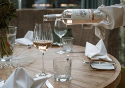 Weinglas mit roséfarbenem Wein auf einem runden Holztisch, daneben ein Glas mit Eiswürfeln und eine Hand gießt Wein aus einer Flasche.