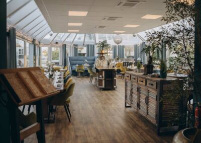 Heller Gastraum mit Holzboden, Tischen mit Stühlen, Pflanzen und großen Fenstern im Dachbereich.