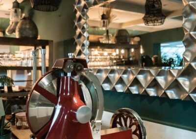 Rote, mechanische Aufschnittmaschine mit großem Drehrad auf Holztisch vor Wandspiegel mit dreidimensionalem, silberfarbenem Rahmen in Restaurant.